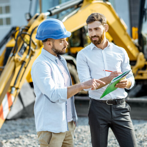 Builder choosing heavy machinery for construction with a sales consultant standing with some documents on the open ground of a shop with special vehicles