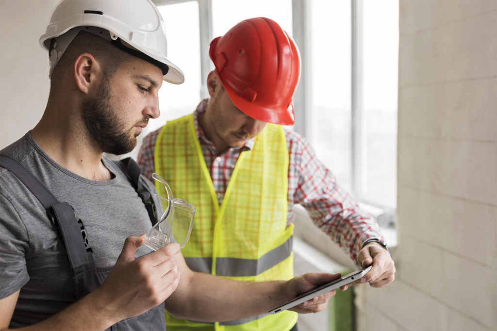 Senior living construction site risk management with two professionals in safety gear reviewing project details on a tablet, assessing progress and safety compliance during ground-up development.
