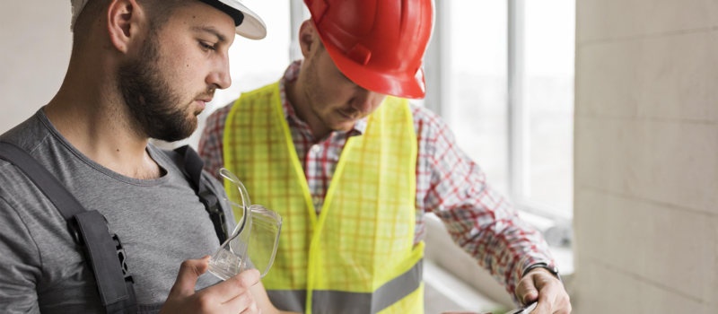 Senior living construction site risk management with two professionals in safety gear reviewing project details on a tablet, assessing progress and safety compliance during ground-up development – zumBrunnen