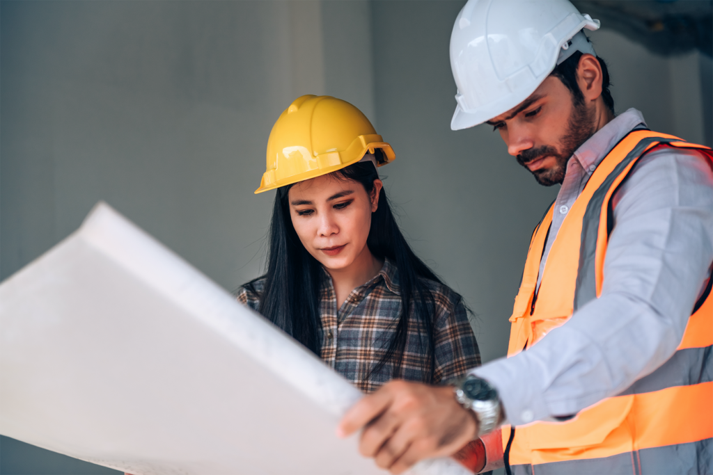 Senior living property condition assessment with two inspectors reviewing building plans on site, wearing safety helmets and evaluating structural details for maintenance and compliance.