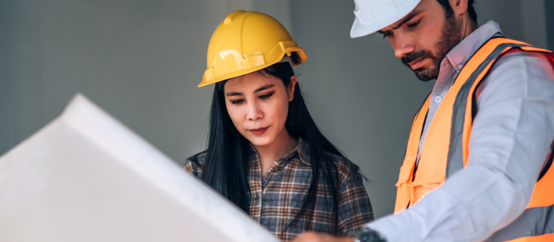 Senior living property condition assessment with two inspectors reviewing building plans on site, wearing safety helmets and evaluating structural details for maintenance and compliance – zumBrunnen